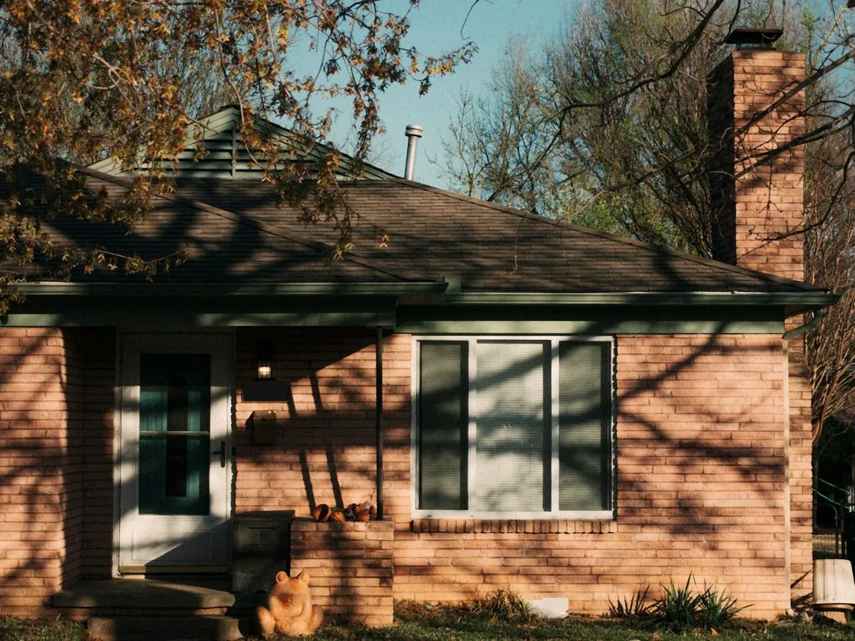Small 1950s brick house in low autumn sun, with a hip roof, green trim, a plain front door and branch shadows across the brick