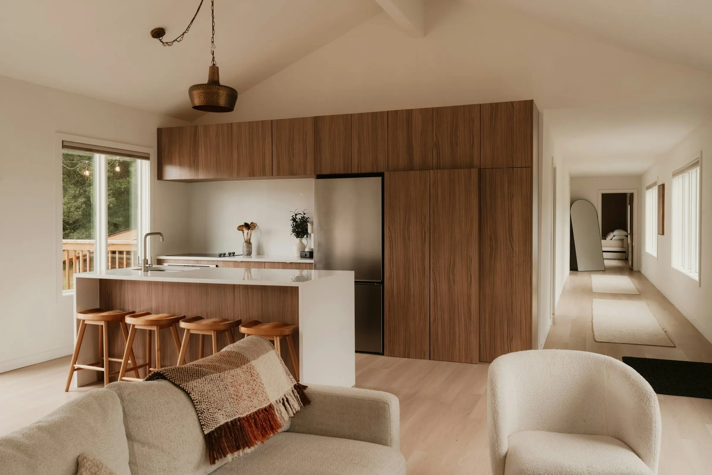 Open kitchen under a vaulted ceiling: a wall of walnut cabinets, a white island with four oak stools and a cream sofa in front